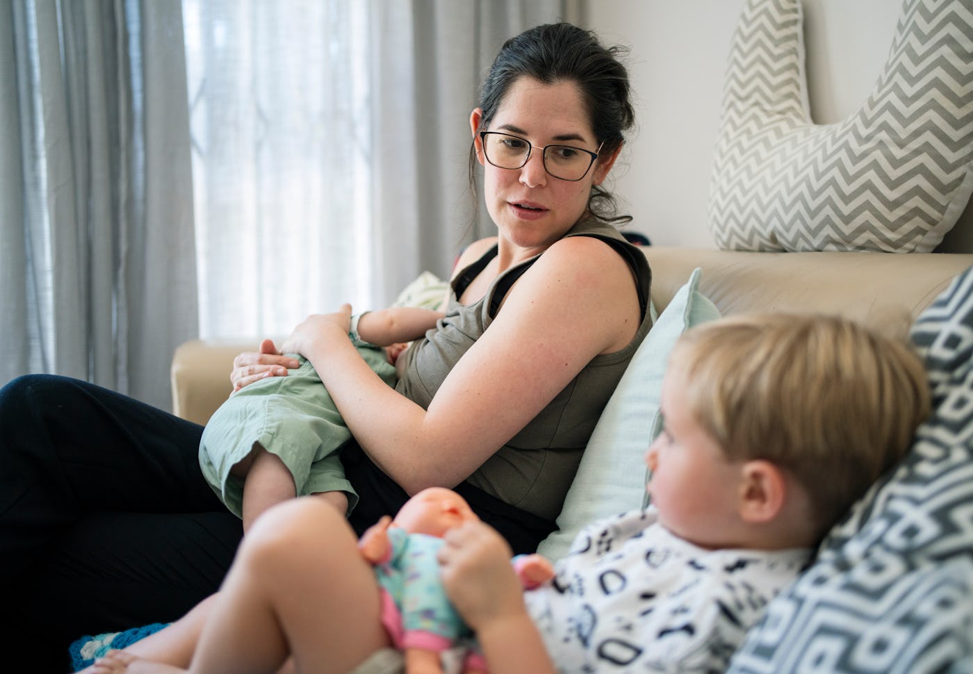 Mother breastfeeding her baby on a sofa at home looking at her toddler son plays with a doll in the living room