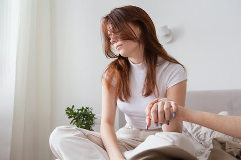 A young woman with long red hair sits on a sofa while receiving emotional support through hand holdi...