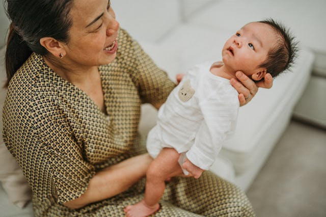A woman in a patterned robe joyfully holds a newborn baby dressed in a white onesie.