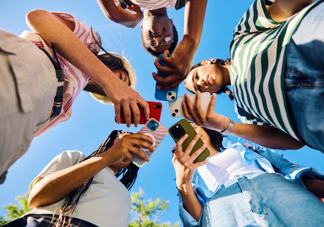 Five high school students are looking down at their smartphones together outdoors