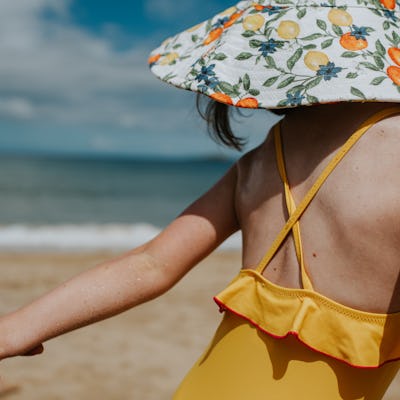 A parent holds a little girls hand and applies spray sunscreen. She wears a swimsuit and sunhat. She glances over her shoulder to watch the sunscreen being rubbed on her arm.