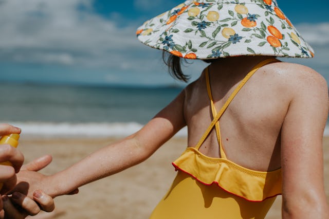 A parent holds a little girls hand and applies spray sunscreen. She wears a swimsuit and sunhat. She...