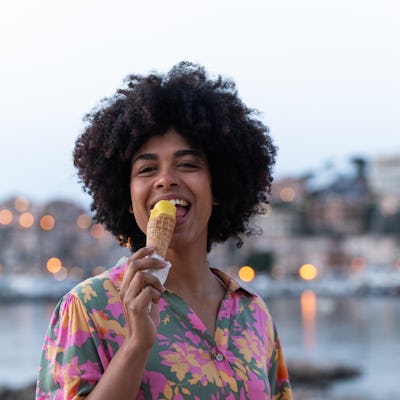 Young woman with curly hair smiles as she enjoys an ice cream cone by the sea at sunset