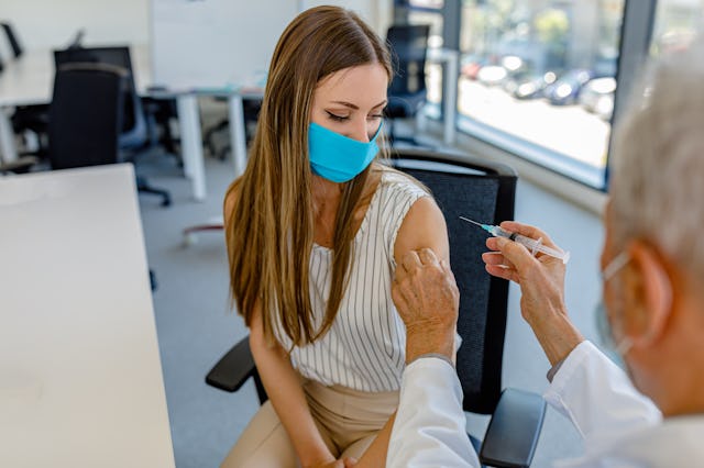 A healthcare professional administers a vaccine to a young woman in a medical setting. Both are wear...