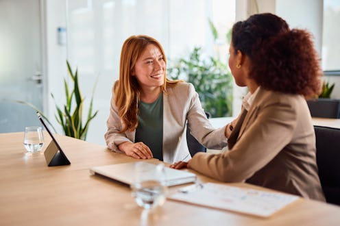 Two businesswomen sitting at a modern office table, engaging in a heartfelt conversation while provi...