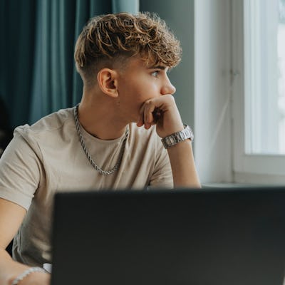 A young man with curly hair sits pensively at a desk, gazing out a window, with a laptop in front of him. A person is seated in the background.