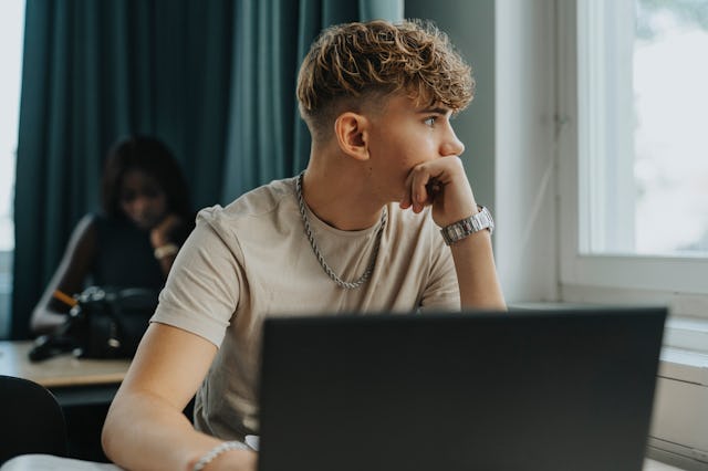 A young man with curly hair sits pensively at a desk, gazing out a window, with a laptop in front of...