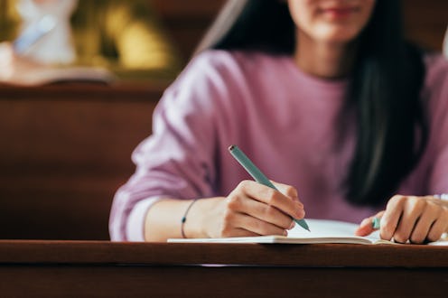 A student focuses on writing study notes in a classroom environment, indicative of concentration and...