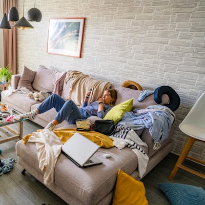 Woman washing the floor of a messy and dirty living room while her lazy daughter is lying on the sofa using phone
