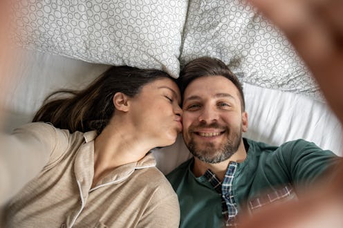 Young couple lying in bed, wearing cozy pajamas, sharing a joyful moment while taking a selfie. Woma...