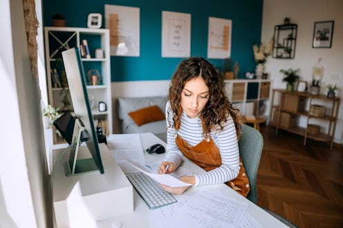 Young female student sits at a desk in her home office, writing in a notebook while studying with de...