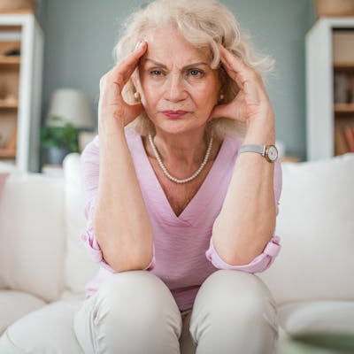 A senior woman is sitting on the sofa in the living room and holding her head because of a severe headache