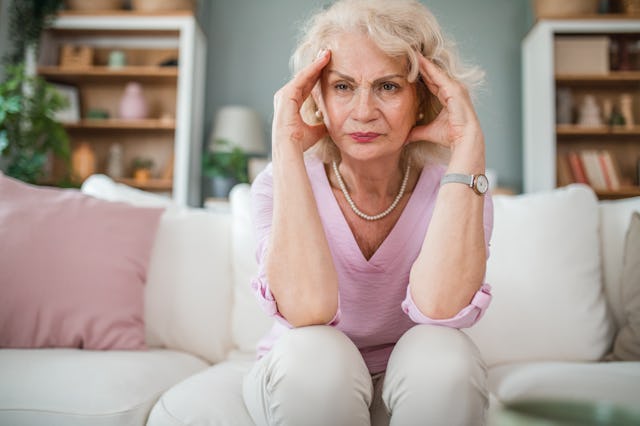 A senior woman is sitting on the sofa in the living room and holding her head because of a severe he...