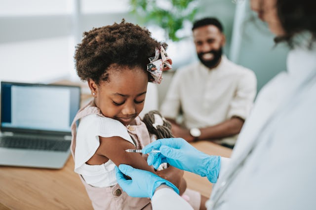 Child being vaccinated, looking down at her arm. Her father is visible in the background.