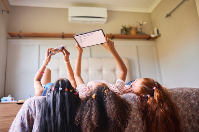 Three girls lie on a bed, one holding a tablet, another a phone, showcasing connected leisure time. ...
