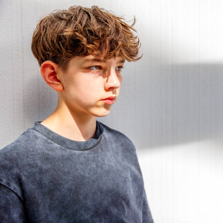 Portrait of a teenage boy with a messed hairstyle leaning on a wall with shadows and looking away th...