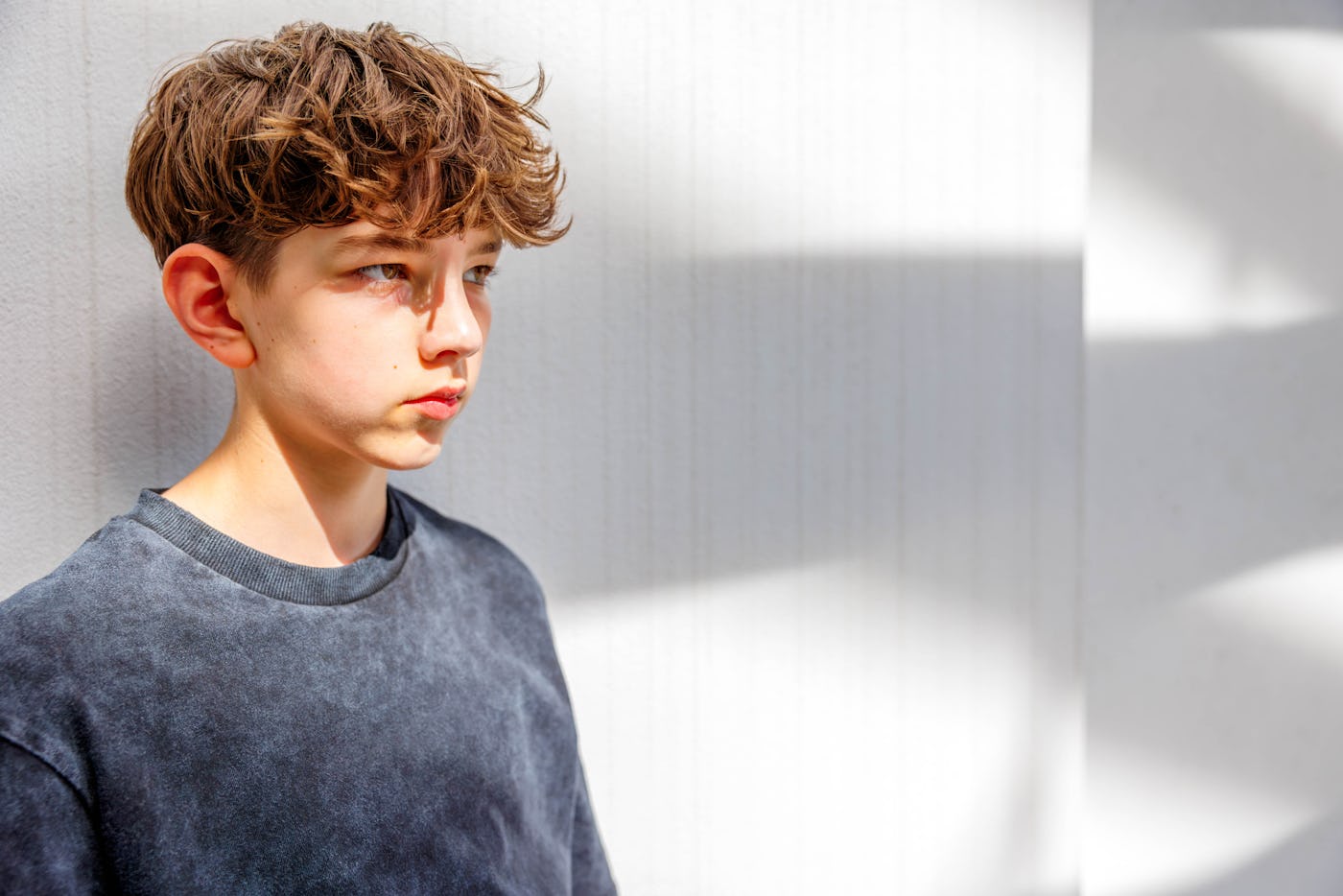 Portrait of a teenage boy with a messed hairstyle leaning on a wall with shadows and looking away thoughtfully