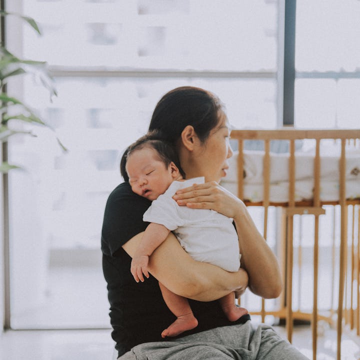 A mother holds her sleeping newborn in a cozy room with natural light, greenery, and a wooden crib n...