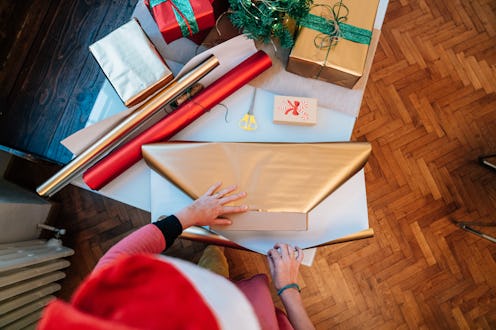 Woman carefully wrapping christmas presents in gold paper, surrounded by colorful rolls and ribbons,...