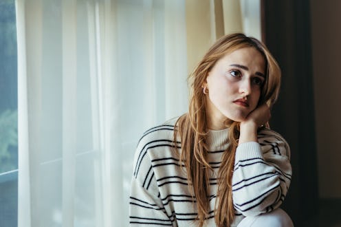 Sad, depressed young woman sitting on the floor by the window. Portrait of lonely, unhappy blond wom...