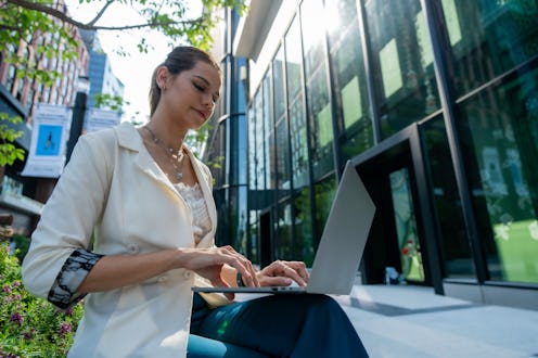 A woman working on laptop outdoors in modern urban setting, surrounded by greenery and glass buildin...