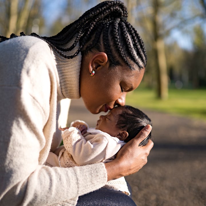 A mother wearing her hair in long braids kisses her baby outside on a park bench.