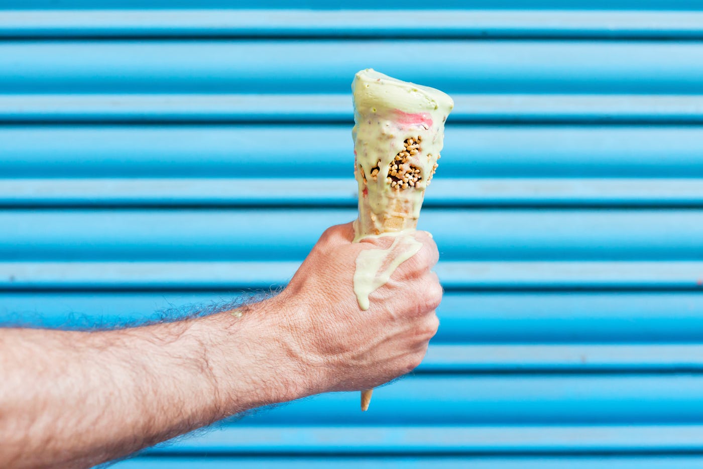 Caucasian adult male hand holding a melting ice cream cone in front of a vibrant blue shutter background. The ice cream is dripping heavily.