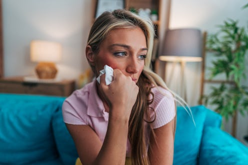 Sad young woman sitting on the sofa at home, wiping tears from her face with a tissue while gazing p...