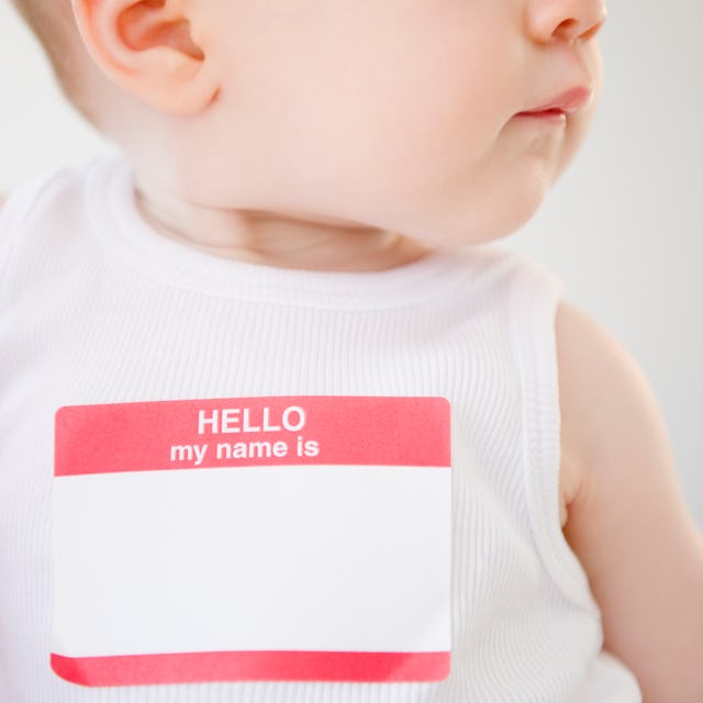 A close-up of a baby wearing a white sleeveless shirt, with a blank name tag reading "HELLO my name ...