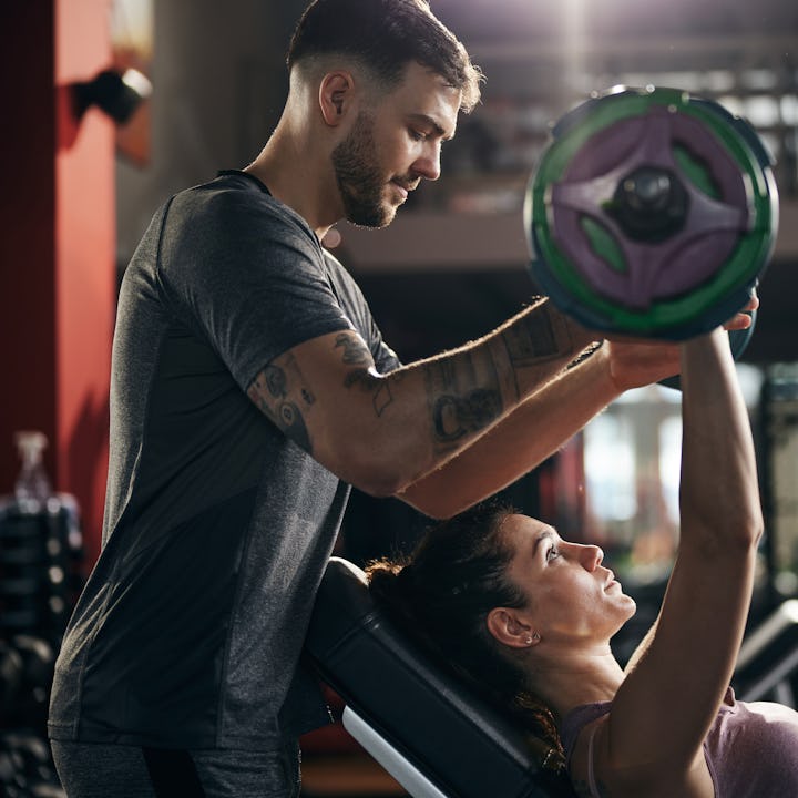 Young man helping his girlfriend during her sports training with barbell in a health club.