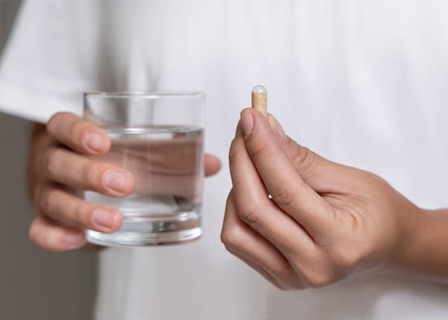 Woman in white t-shirt holding beige capsule in fingers and water glass in hand closeup. Female or m...