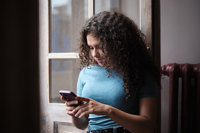 A young woman with curly hair stands by a window, looking intently at her smartphone. Soft natural l...