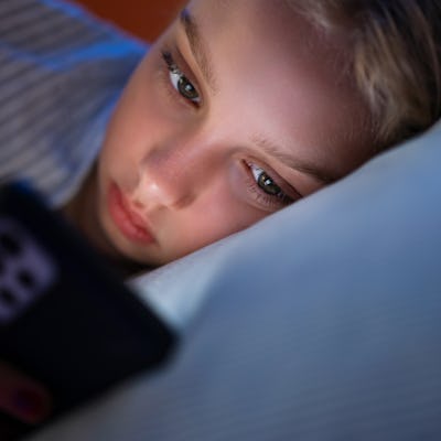 Close-up of a young girl lying in bed at night, illuminated by the light of a smartphone, focused on the screen.