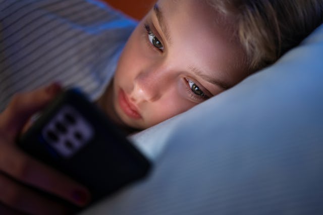 Close-up of a young girl lying in bed at night, illuminated by the light of a smartphone, focused on...
