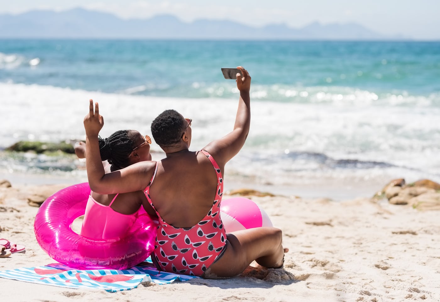 Rear view shot of two sisters sitting on the beach and taking selfie