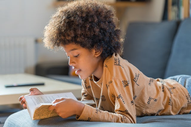 Side view of an African American boy reading a book lying on the couch at home
