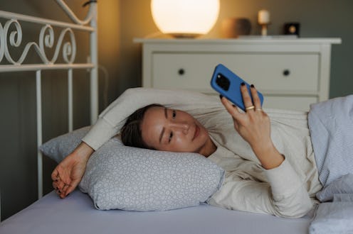 Korean woman relaxing in bed while browsing on smart phone, illuminated by soft ambient lighting.