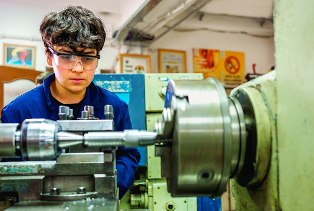 A young male trainee in safety glasses operates a Lathe machine in a technical training workshop. Co...