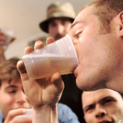 A group of young men are gathered around, with one man in the foreground drinking from a clear plastic cup. The atmosphere feels casual and lively.