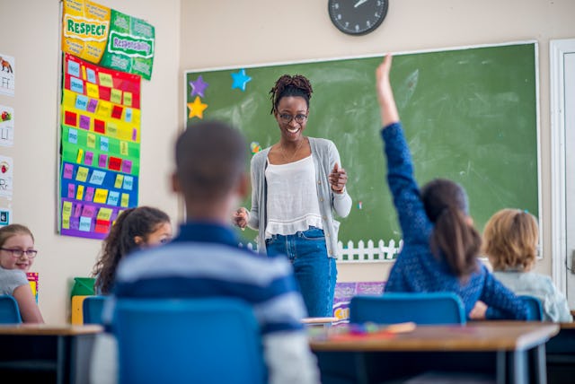 A teacher is standing at the front of her elementary school classroom. One girl is raising her hand ...