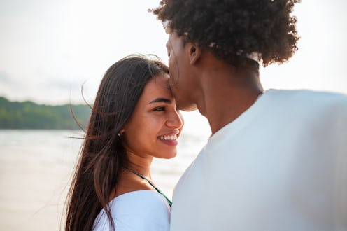 A Hispanic woman and a Black man share a tender kiss, dressed in casual beachwear, with the serene b...