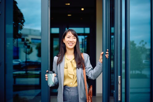 Young businesswoman is arriving at the office, opening the glass door and holding a disposable coffe...