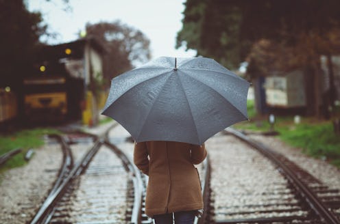 A woman under a grey umbrella strolls along wet railway tracks amidst a rainy setting. The surroundi...