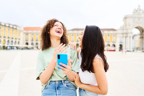 two female tourists standing in a square laughing together at something funny they saw on the phone,...