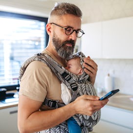 Father scrolling on smartphone while newborn child sleeping in baby carrier. Concept of paternity le...