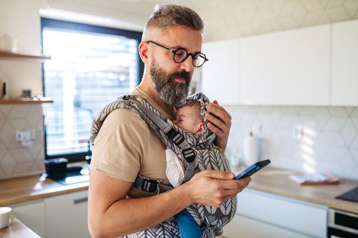 Father scrolling on smartphone while newborn child sleeping in baby carrier. Concept of paternity le...
