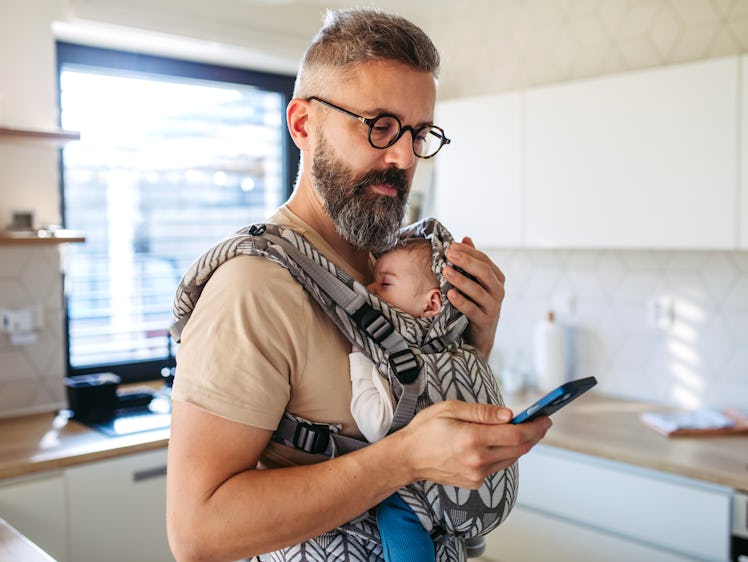 Father scrolling on smartphone while newborn child sleeping in baby carrier. Concept of paternity le...