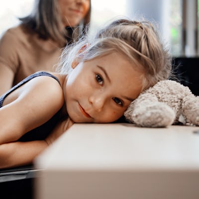 Little girl rests head on table with teddy bear and looking at camera with her mother sitting behind in doctor office