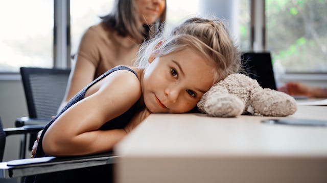 Little girl rests head on table with teddy bear and looking at camera with her mother sitting behind...
