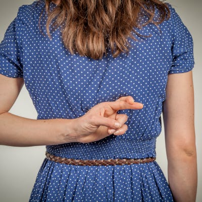 A woman stands with her back to the camera, wearing a blue polka dot dress. Her right hand is crossing her fingers behind her back, suggesting dishonesty.